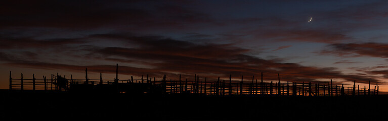 A panoramic image of a ranch corral silhouetted at dusk with some orange light lingering in the sky painting the clouds while the crescent moon hangs above the horizon to the right.