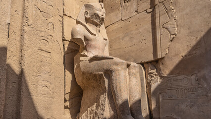 A damaged ancient sculpture of a seated pharaoh against the background of stone walls with...