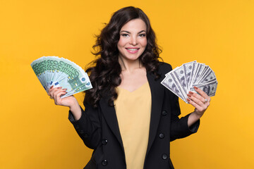Young cheerful wealthy woman holding two cash fans of hundred euro and dollar banknotes of money,...