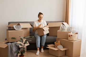 Portrait of smiling positive woman wearing white casual T-shirt and jeans, sitting on sofa, surrounded with boxes during moving, unpacking personal pile, expressing happiness.