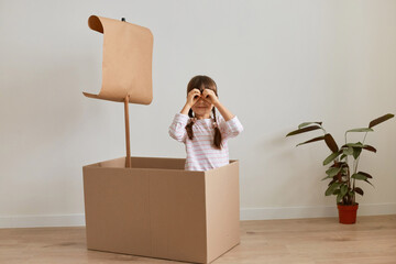 Indoor shot of cute funny little girl sitting in cardboard box with paper flag, playing ship...
