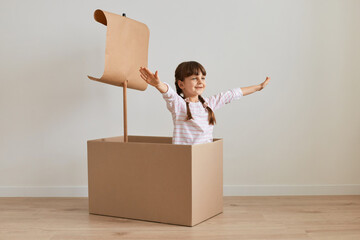 Indoor shot of positive playful little girl sitting in cardboard box with paper flag, playing ship...