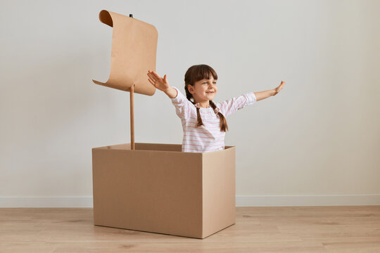 Indoor shot of positive playful little girl sitting in cardboard box with paper flag, playing ship captain, posing with raised arms and happy expression, pretending she is in the sea.