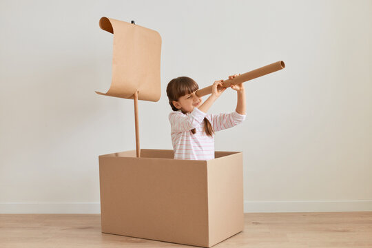 Portrait of positive funny little girl sitting in cardboard box with paper flag, playing ship captain, pretending she is in the sea, holding big spotting scope, looking far.