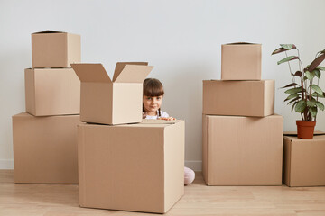 Indoor shot of shy little dark haired girl sitting on floor surrounded with cardboard boxes with personal pile, hiding behind carton boxes, looking at camera.