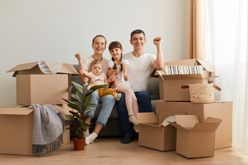 Happy family sitting on cough in a new apartment, celebrating relocation, clenched fists, moving to a new big light house, being surrounded with cardboard packages.