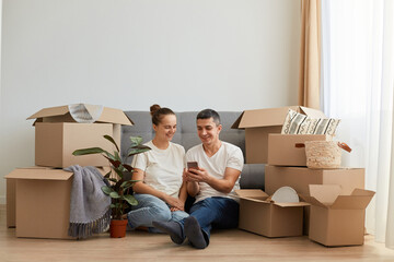 Happy husband and wife move in new bought house, buy furniture online, use modern technology, man surfing internet via laptop and talking over phone, sitting on floor near sofa.