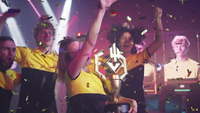 Cheerful blond young woman gamer winner with golden trophy clenching fist and jumping while celebrating victory in gaming championship with teammates