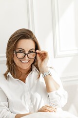 Portrait of an attractive smiling middle-aged woman in stylish glasses. Beautiful positive woman in a white cotton shirt. Vertical shot