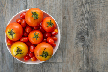 Top view closeup studio shot of two healthy fresh delicious red and cherry tomatoes vegetable species in white plastic bowl placing on old wooden table background for advertisement with copy space
