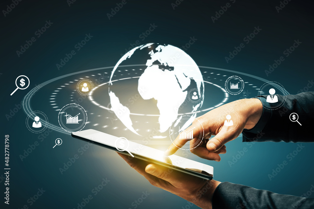 Poster close up of businessman hand pointing at tablet with glowing social globe hologram on blue backgroun