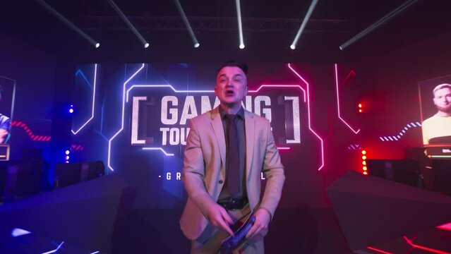 Excited adult announcer speaking to camera at start of professional esports competition on glowing stage with digital screen and neon illumination