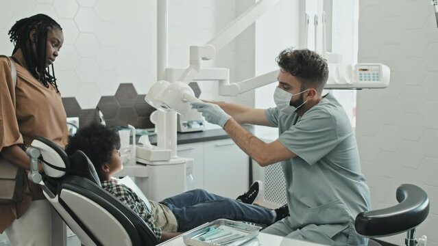 Medium long of male Caucasian doctor wearing scrubs, gloves and face mask in his office, using mouth mirror on Black elementary-aged boy sitting in dentist chair, his mother standing by side