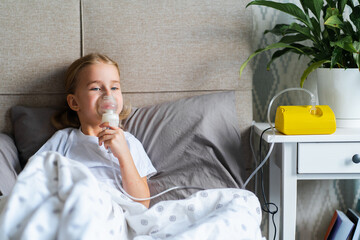 Sick little girl making inhalation with nebulizer to reduce coughing, lying in bed at home, child taking medication while breathing in through face mask. Bronchitis and asthma treatments for children - obrazy, fototapety, plakaty