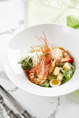 Classic Caesar salad with shrimps in white plate on stone table. Italian menu in restaurant. Seafood caesar on rustic style. Trendy salad with grilled prawns, green leaves, eggs, croutons and cheese.
