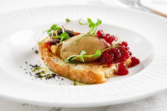 Chicken liver pate on crispy bread with red currant jam and micro green. Bruschetta with chicken pate in white plate. Delicious appetizer - foie gras on toast. Liver pate with jam in rustic style.