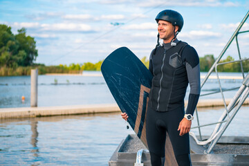 Smiling wakeboarder in wetsuit and helmet with board standing on pier
