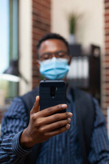 Closeup of entrepreneur holding smartphone in video call conference wearing covid protective mask and wireless earbuds. Detail view of startup employee smart phone in online interview.