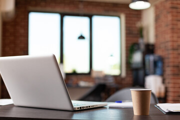 Close up of laptop computer and coffee cup on desk in empty business office. Nobody in modern startup space with electronic equipment and beverage in plastic cup on wooden table.