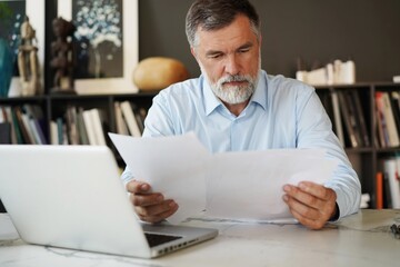 Serious mature male employee sits at the table in modern office, works with documents