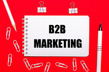 On a red background, a white pen, white paper clips, a white pencil and a notebook with the text B2B MARKETING. View from above