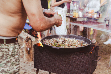 A man cooks food on a campfire. Large frying pan with champignons and herbs.