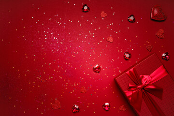 Valentine's Day gift in a red box tied with a red ribbon and red crystals, heart-shaped rubies, rhinestones, glitter on a red background with copy space.