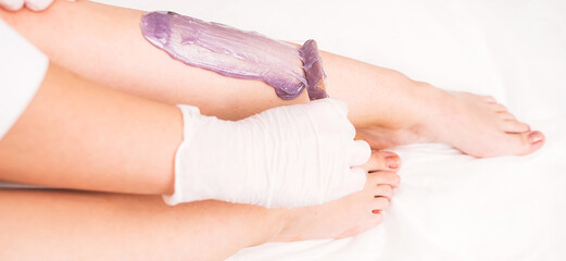 Cosmetologist in white gloves applies warm wax using spatula to a woman leg to make hair removal procedure on legs in beauty salon.