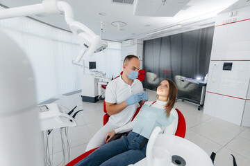 A young dentist specialist examines and treats the teeth of a beautiful woman in modern white...