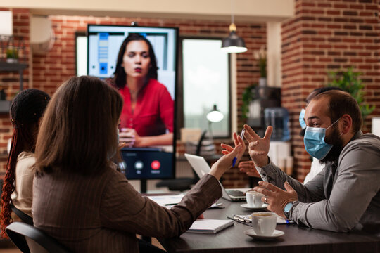 Diverse businesspeople with medical face masks discussing marketing statistics planning company strategy with remote manager in startup business office. Online videocall meeting conference on monitor