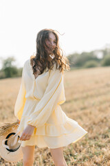 Happy young girl running through a yellow field on a summer day. girl in yellow dress and straw hat with a bouquet of wheat. sheared golden wheat spikelets. Agricultural texture