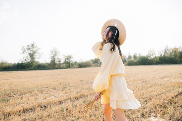 Caucasian young brunette walking through a wheat field. girl in yellow summer dress and straw hat. sheared golden ears of wheat, rye. Close-up portrait of a beautiful girl with brown eyes. 