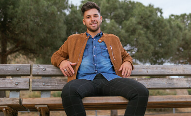 Fototapeta premium Shot of a handsome guy in casual clothes sitting on a bench of a viewpoint and smiling while looking at the camera