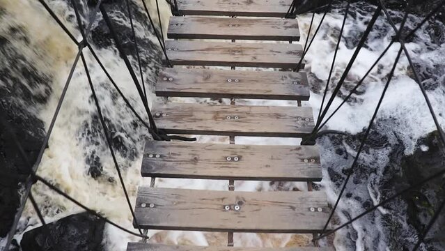 Movement on suspension swinging bridge made of ropes and wood boards over rocky river with waterfall. POV video shooting.