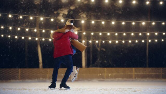 Romantic Winter Snowy Evening: Ice Skating Couple Having Fun on Ice Rink, Spin, Dance, Jump. Pair Skating Outdoors. Portrait of Young People, Boyfriend and Girlfriend in Love. Slow Motion Wide shot