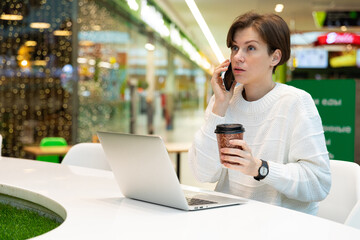 Young attractive woman sitting at a shopping center at a table and working at a computer laptop, using mobile phone, drinking coffee. Cafe interior. Freelance and business concept