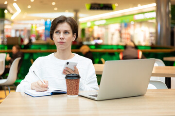 Photo of young attractive brunette woman sitting at a shopping center at a table with coffee paper cup and working at a computer laptop, using mobile phone. Freelance and business concept