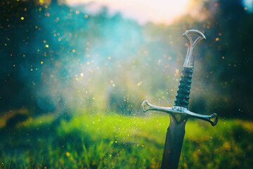 mysterious and magical photo of silver sword over England woods or field landscape with light flare. Medieval period concept.