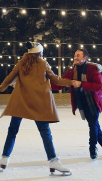 Vertical Video. Romantic Winter Evening: Ice Skating Couple Having Fun on Ice Rink. Beautiful Snow Falls on Pair Skating Boyfrined and Girlfriend in Love, Dance, Hold Hands, Embrace, Figure Skate