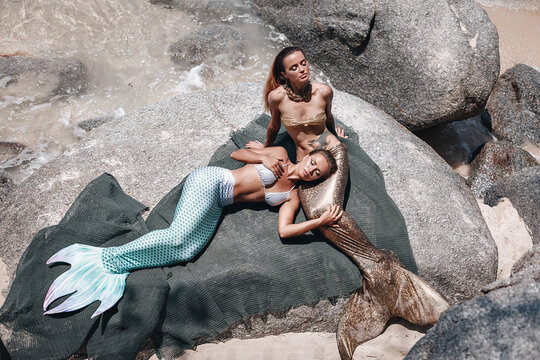 two gorgeous mermaids with long tails are lying on the rocks and posing on the beach, sunbathing under the hot sun