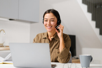 Young cheerful woman using headset for online talking sitting at the desk at home office. Pleasant...