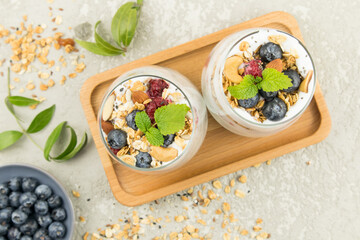 Fototapeta na wymiar two glass glasses with a delicious healthy breakfast of granola with yogurt, nuts, berries and mint stand on a wooden tray. top view. gray background.