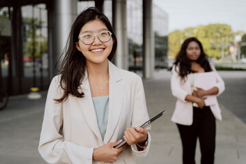 The end of work in the office, co-workers go outside from company building, young business people woman with Asian features in glasses with brown hair smiles holds tablet in hand satisfied with career