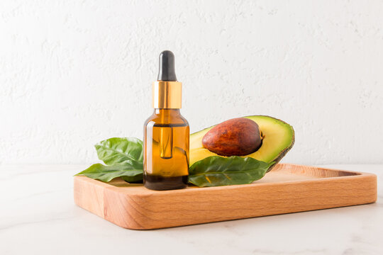 A cosmetic glass bottle of organic avocado oil stands on a wooden tray with ripe fruit and leaves opposite a white brick wall. natural cosmetics.