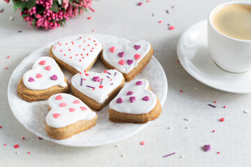 Gingerbread covered with white icing and sprinkling on a light saucer with a cup of espresso and flowers in the background. The concept of celebrating Valentine's Day. Horizontal orientation.