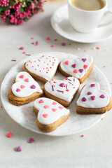 Gingerbread covered with white icing and sprinkling on a light saucer with a cup of espresso and flowers in the background. The concept of celebrating Valentine's Day. Vertical orientation.