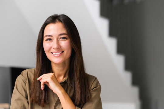 Portrait of charming elegant asian woman with hand under chin smiling at the camera, sitting in the modern house, wearing domestic clothes. Stock photo