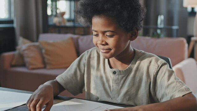 Tilting up of ten-year-old Black boy drawing at desk at home, eating sweets at daytime