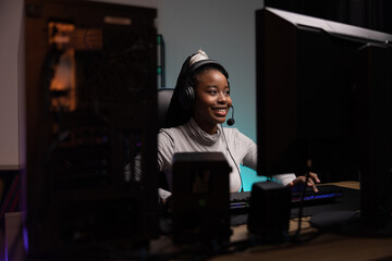 Shot from behind a computer monitor of a smiling pro gamer. Girl has headset to play goes round in game, wins over other participants Dark room at night lit by led lights