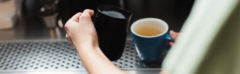 Cropped view of barista holding milk jug and cup in cafe, banner.